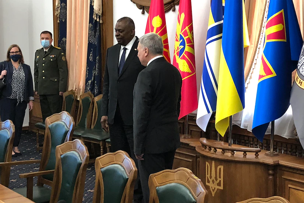 Two men speak inside a room with flags behind them.