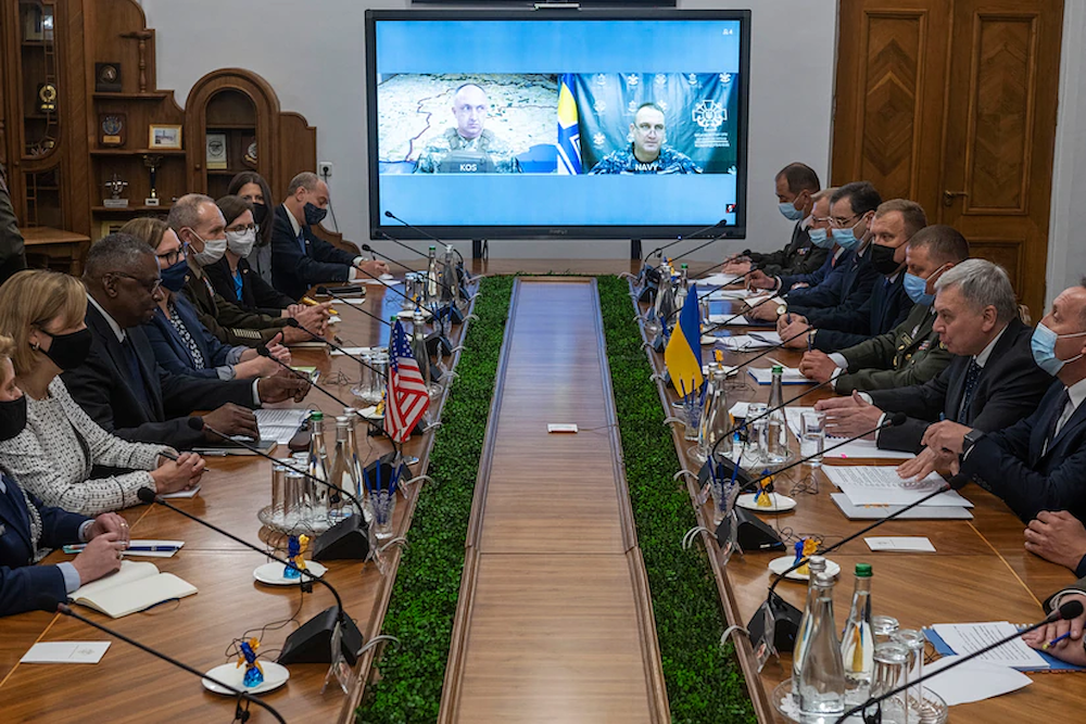 Two delegations sit at a long table with others attending via teleconference.